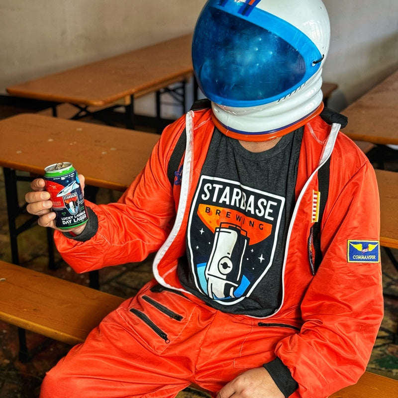 An astronaut wearing a Starbase Brewing shirt drinking a Starbase beer.